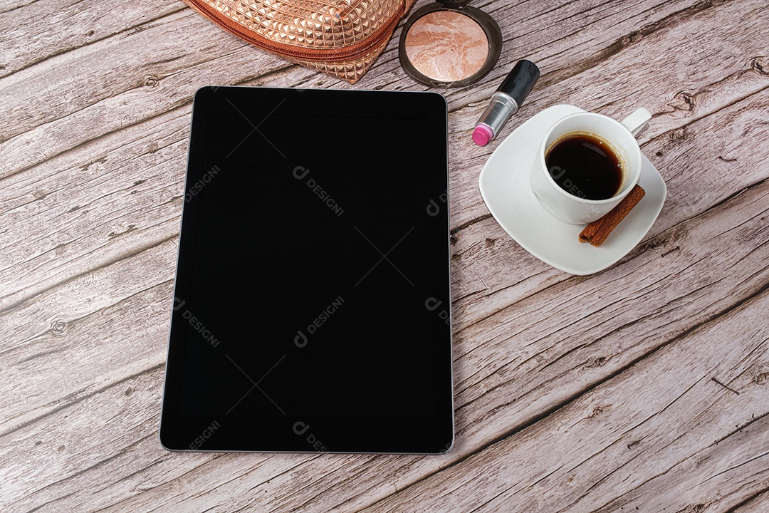 Tablet na mesa cercado por smartphone, batom, pó compacto, xícara de café e maquiagem