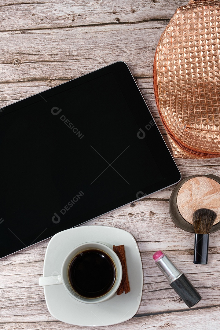 Tablet na mesa cercado por smartphone, batom, pó compacto, xícara de café e maquiagem