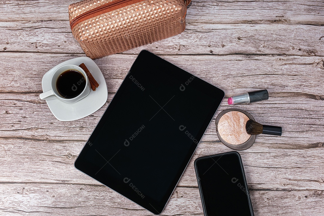 Tablet na mesa cercado por smartphone, batom, pó compacto, xícara de café e maquiagem