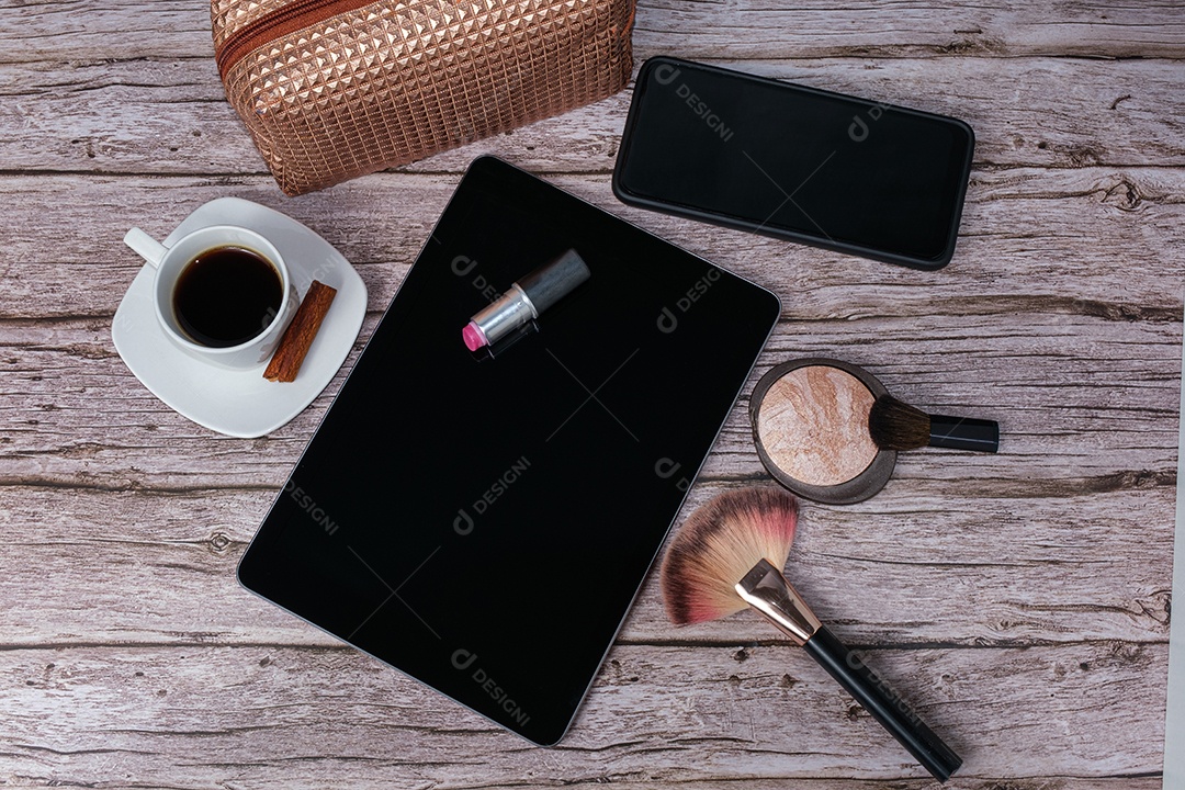 Batom no tablet cercado por xícara de café, bolsa de maquiagem, smartphone, pó e pincel.
