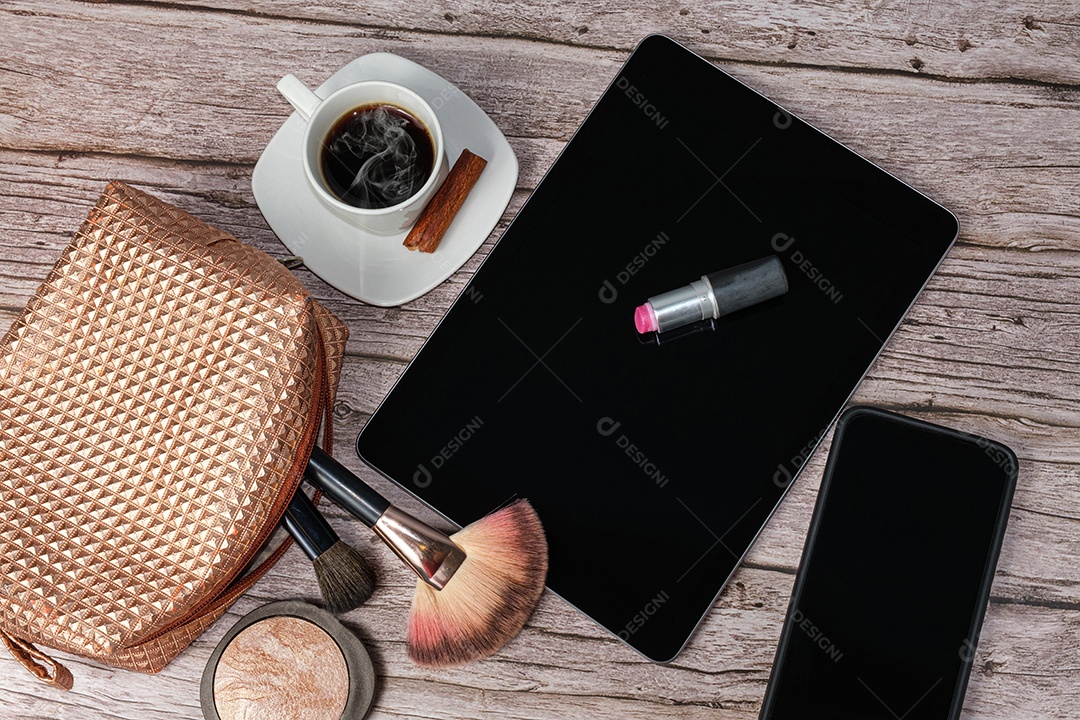 Batom no tablet cercado por xícara de café, bolsa de maquiagem, smartphone, pó e pincéis.