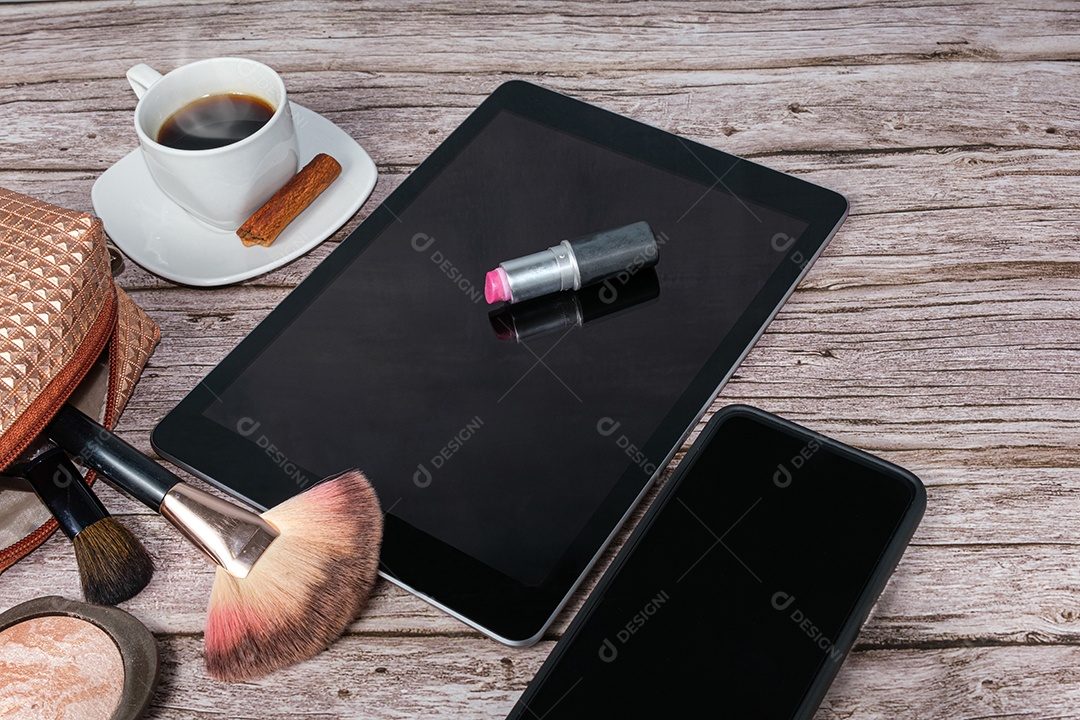 Batom no tablet cercado por xícara de café, bolsa de maquiagem, smartphone, pó e pincéis.