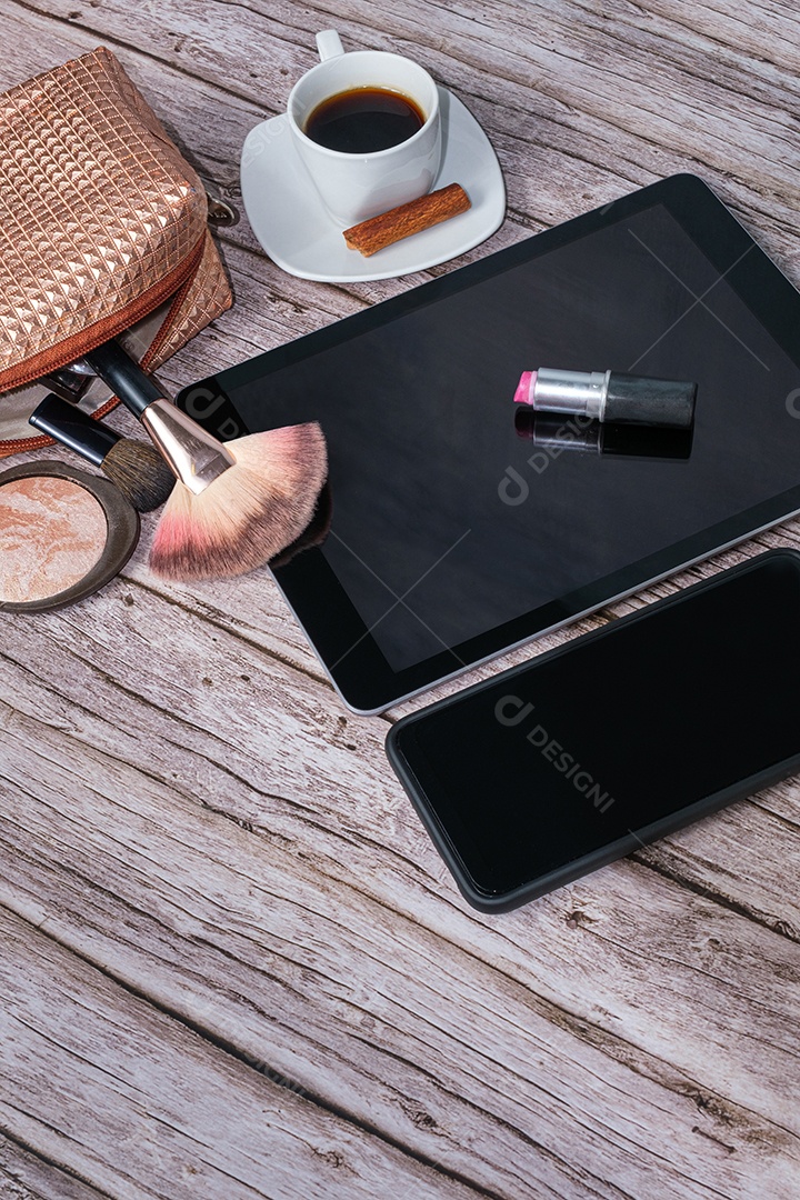 Batom no tablet cercado por xícara de café, bolsa de maquiagem, smartphone, pó e pincéis.