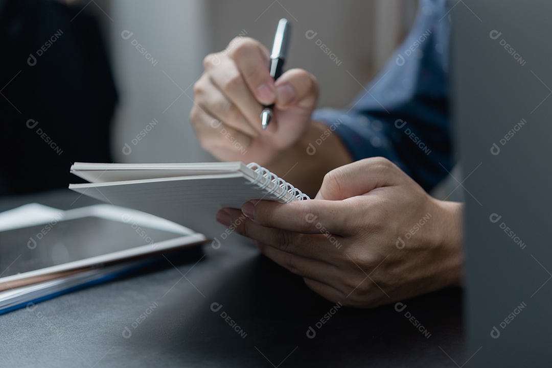 Mão masculina escrevendo no caderno com caneta no computador portátil no escritório moderno.