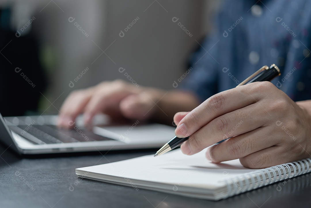 Mão masculina escrevendo no caderno com caneta no computador portátil no escritório moderno.