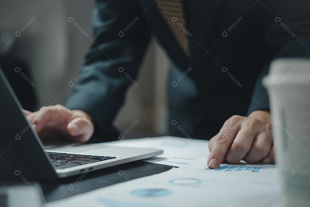 Mão de empresário apontando estatística de análise de relatório de gráfico de papelada e gráfico financeiro de documento com computador portátil no escritório.