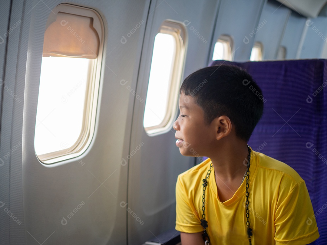 Um menino asiático senta-se e sorri e olha pela janela de um avião.