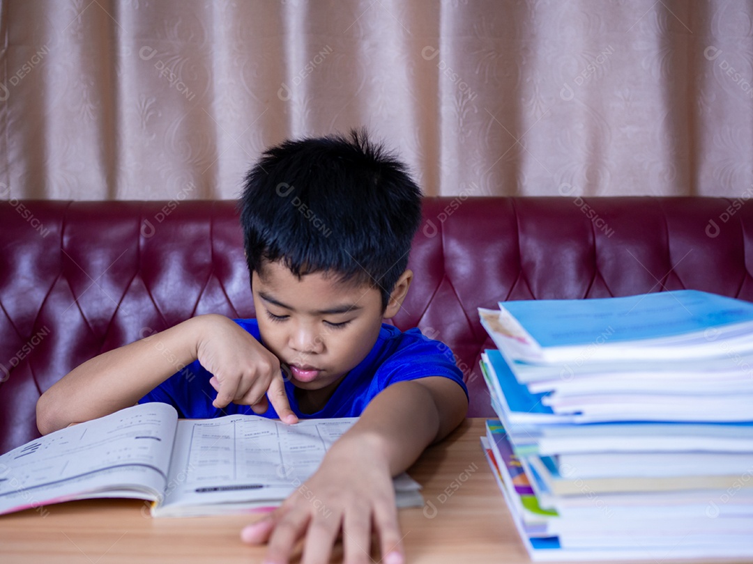 Menino fazendo lição de casa e lendo em uma mesa de madeira com uma pilha de livros.