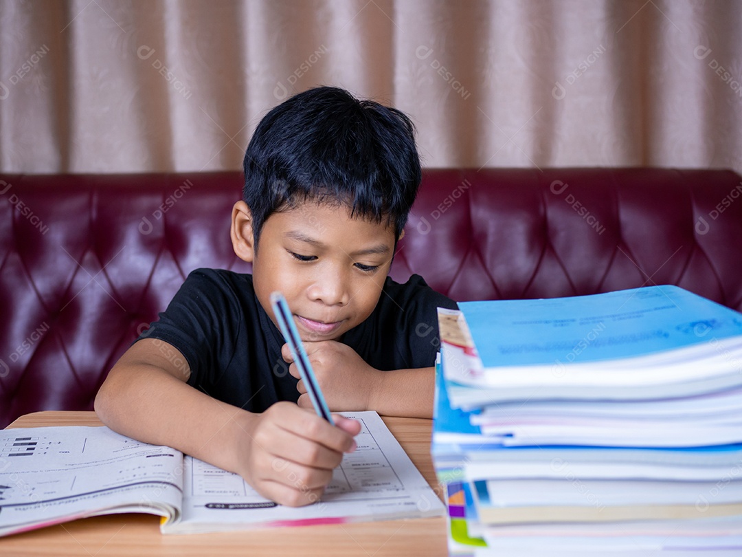 Menino fazendo lição de casa e lendo em uma mesa de madeira com uma pilha de livros.