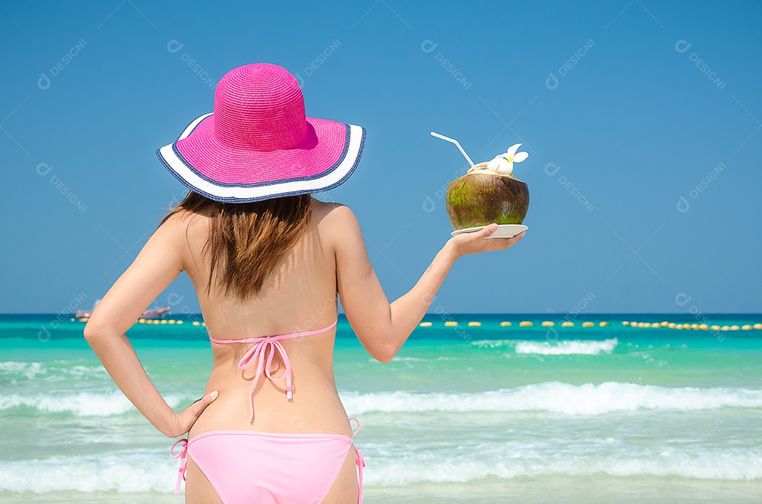 Mulher asiática usa um biquíni rosa na praia e segura coco
