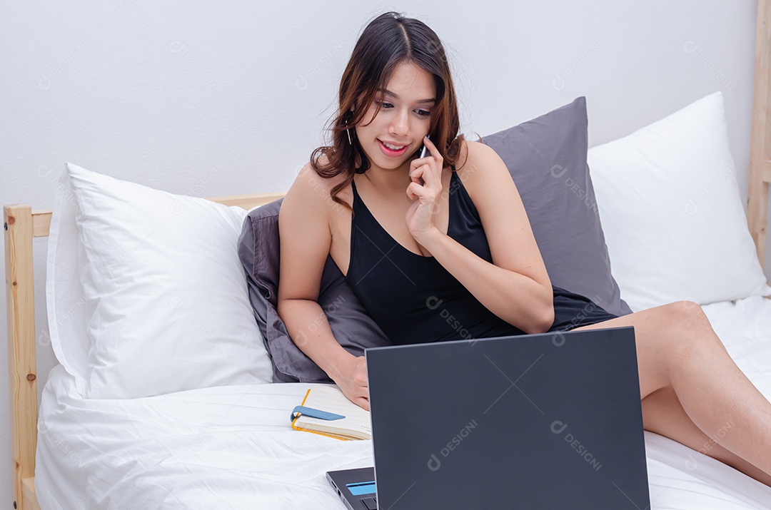 Linda mulher asiática está relaxando usando celular e notebook computador na cama.