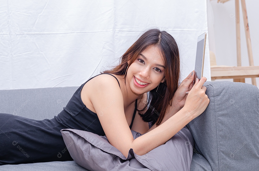 Linda mulher asiática está relaxando e usa tablet no sofá, tablet de uso feminino para mensagens de texto, leitura, assistir a vídeos e fazer compras online.