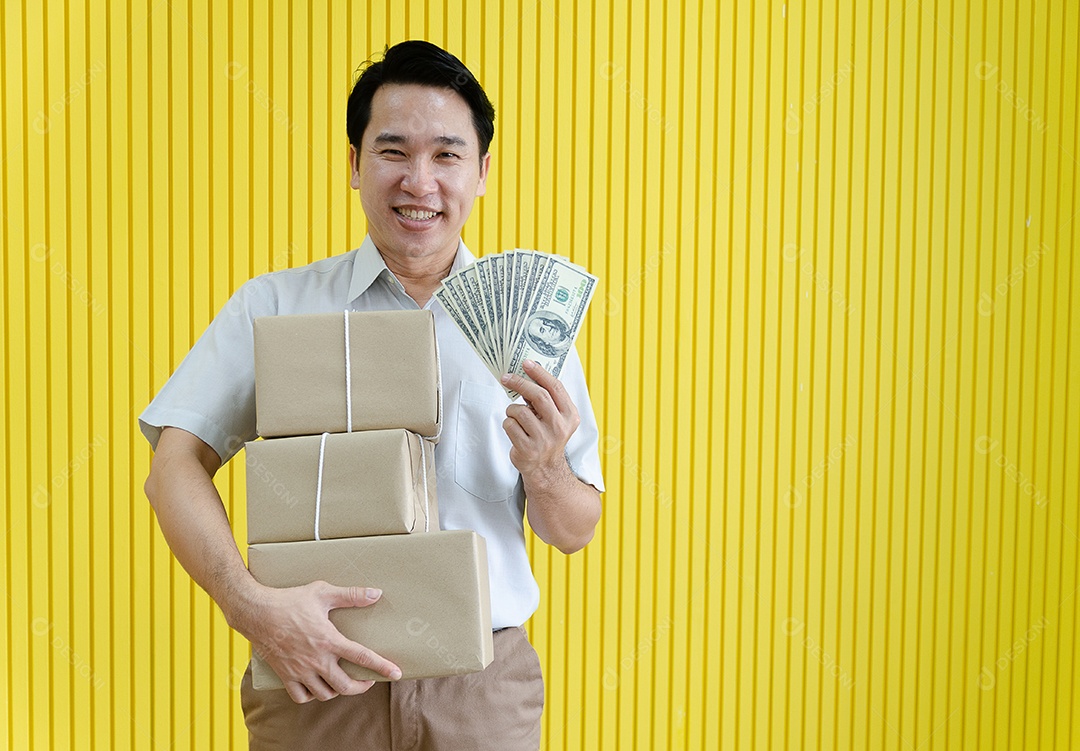 Pequeno empresário asiático feliz segurando dólar e preparando mercadorias para clientes, fundo amarelo.
