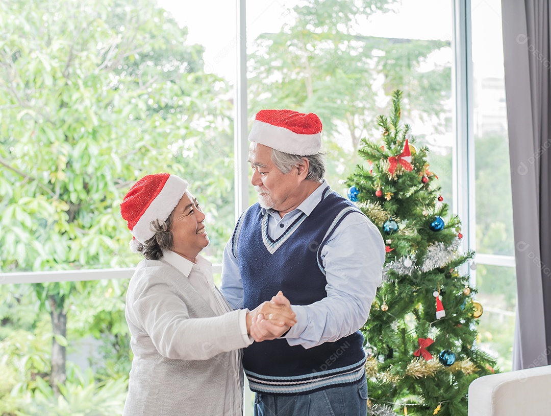 Casal senhor, marido e mulher usando chapéus de natal e dançando, árvore de fundo.