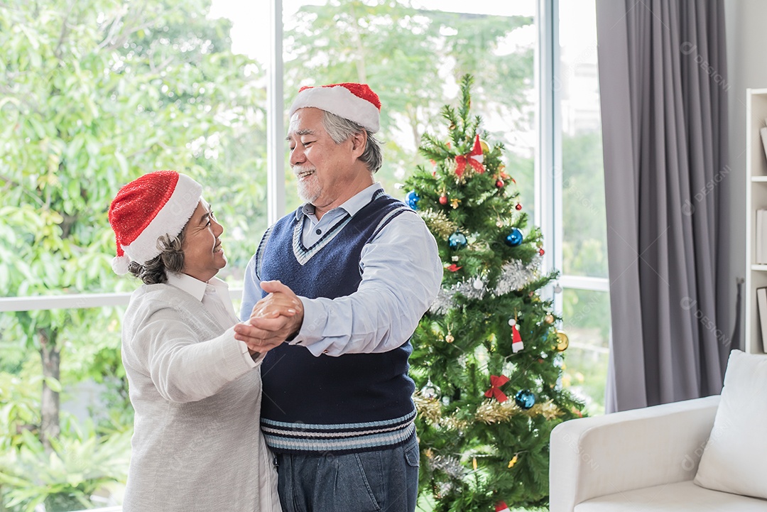 Casal senhor, marido e mulher usando chapéus de natal e dançando, árvore de fundo.