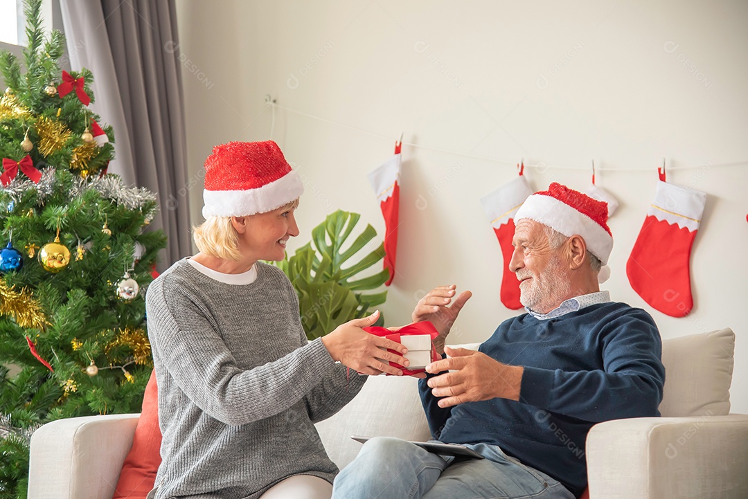 Marido mais velho dá uma caixa de presente para sua esposa sentado no sofá e usando um chapéu