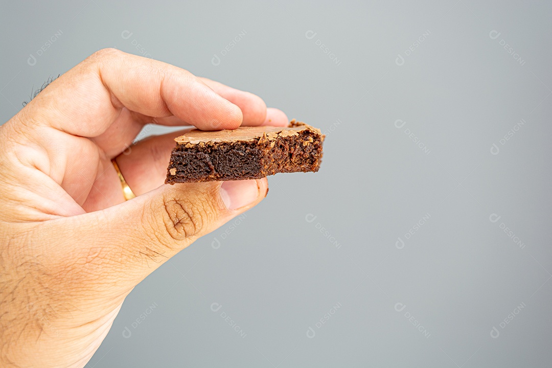 mão masculina segurando brownie de chocolate mordido em fundo cinza