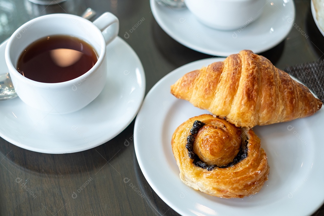 Pão, croissant em um prato branco com um copo de chá na mesa do café.