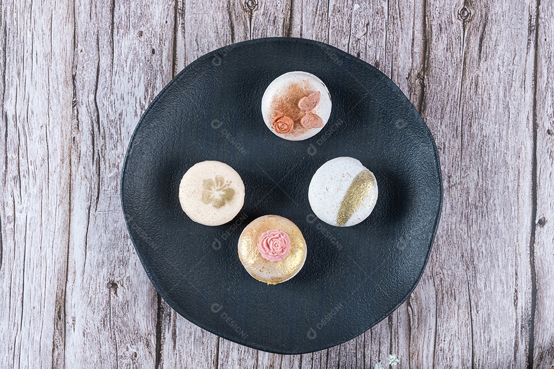 Quatro macarons, decorados com tinta comestível e fondant, em um prato preto.