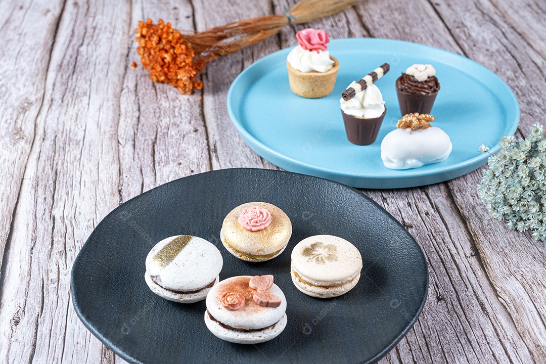 Macarons decorados com tinta comestível e fondant. Ao fundo, doces variados com chantilly e nozes.