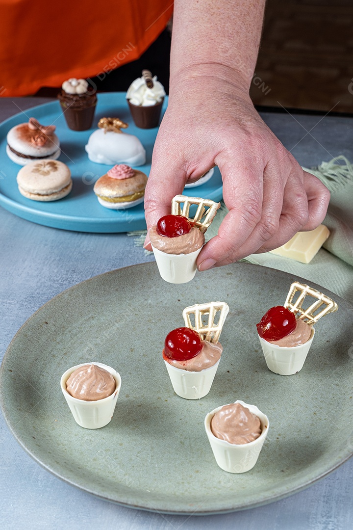 Confeiteiro pegando um pequeno copo de chocolate branco cheio de mousse de chocolate ao leite e cereja.