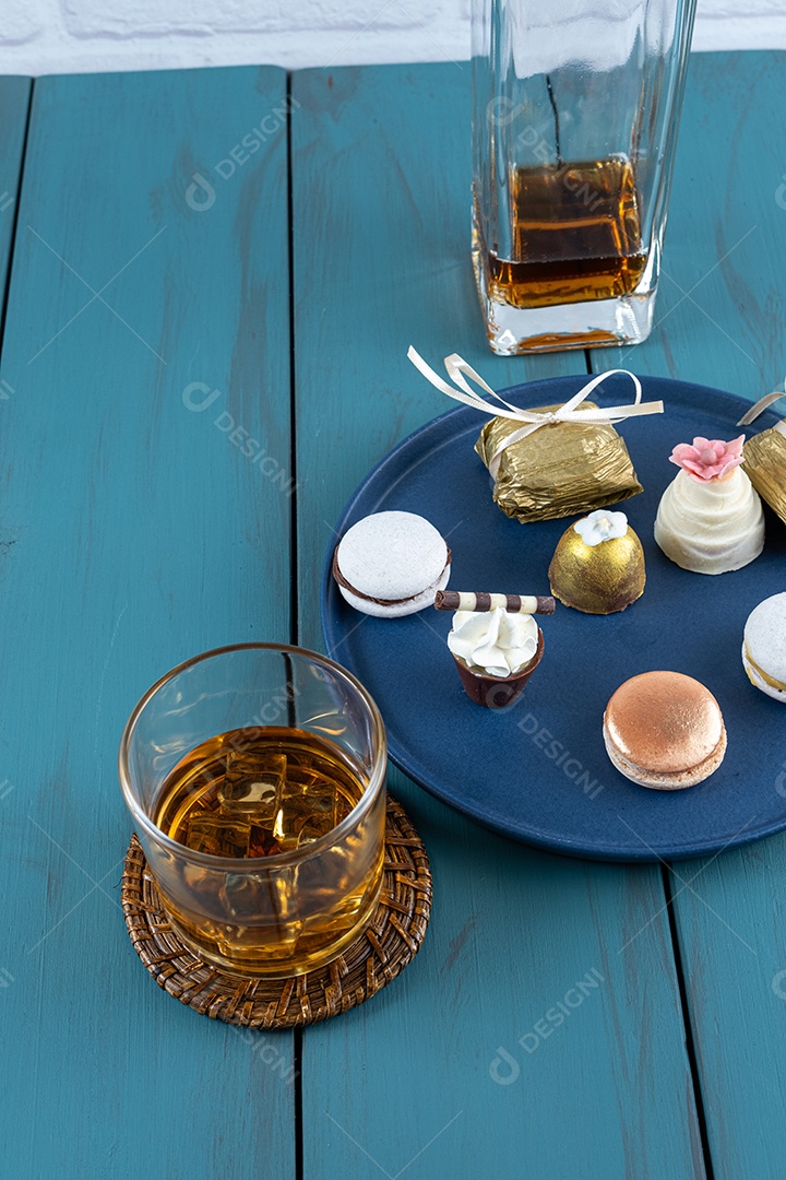 Copo pequeno de chocolate com chantilly, ao lado de macarons, camafeu de noz, trufa, garrafa e copo de uísque.