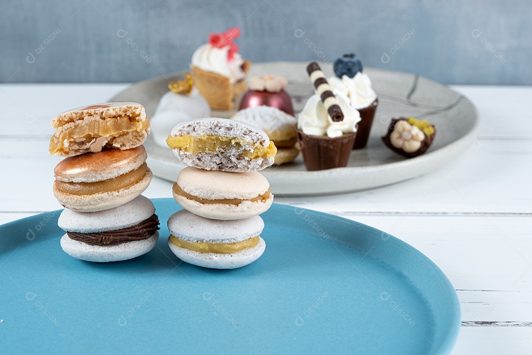 Bolo de chocolate branco pequeno ao lado de doces variados. No primeiro plano desfocado, macarons empilhados.