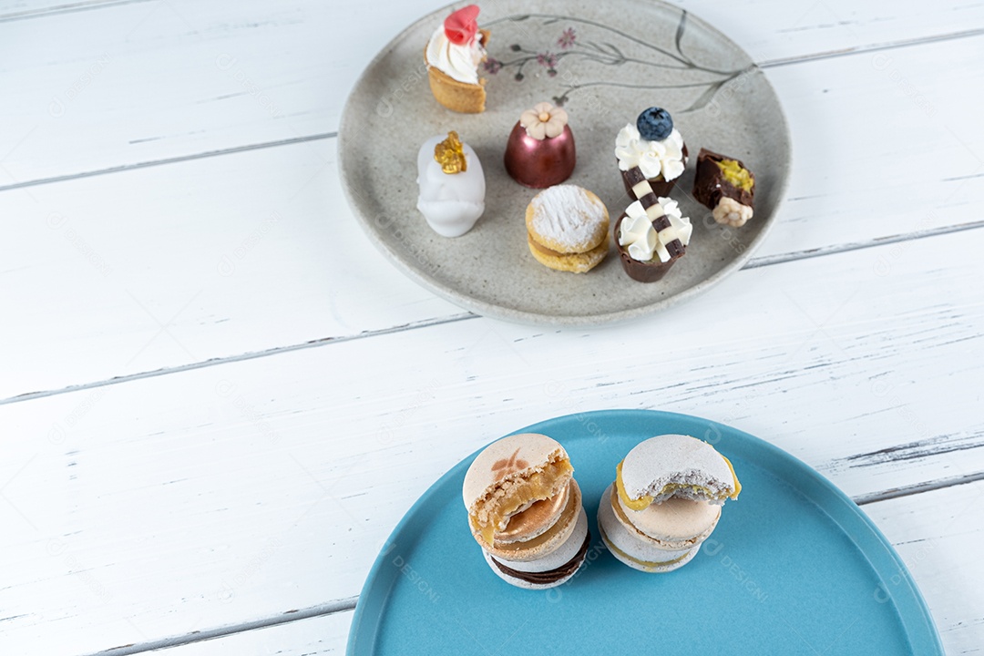 Bolo de chocolate branco pequeno ao lado de doces variados. No primeiro plano desfocado, macarons empilhados.
