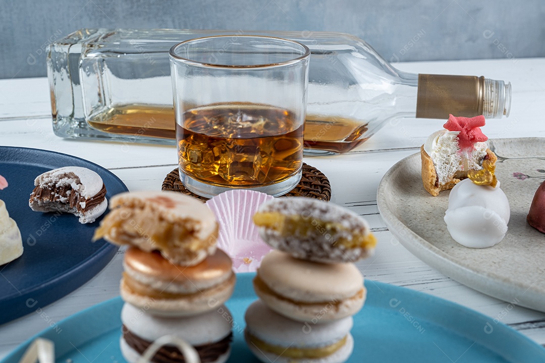 Copo de uísque com gelo cercado por doces de casamento variados. No primeiro plano desfocado, macarons empilhados.