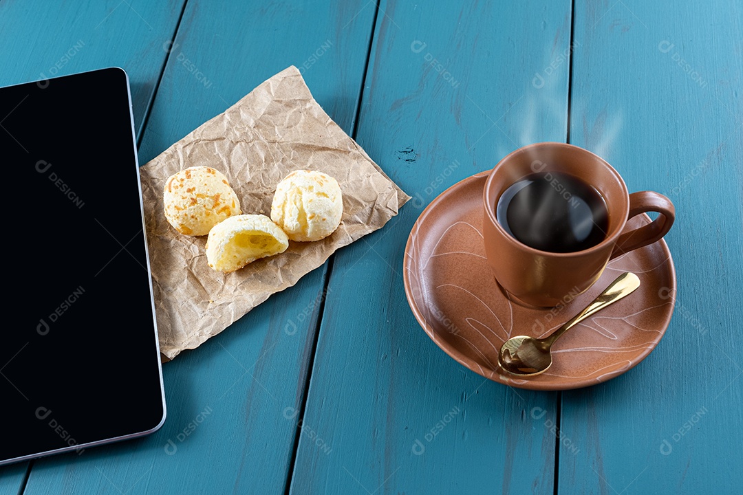 Closeup de pães de queijo brasileiros em um papel pardo, ao lado de tablet, xícara de vista.
