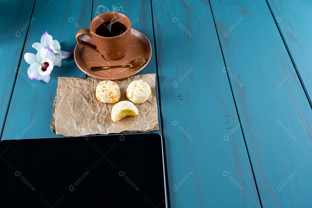 Closeup de pães de queijo brasileiros em um papel pardo, ao lado de tablet, xícara de vista.