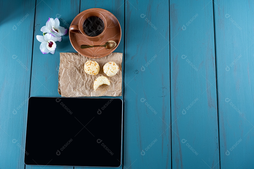 Closeup de pães de queijo brasileiros em um papel pardo, ao lado de tablet, xícara de vista.