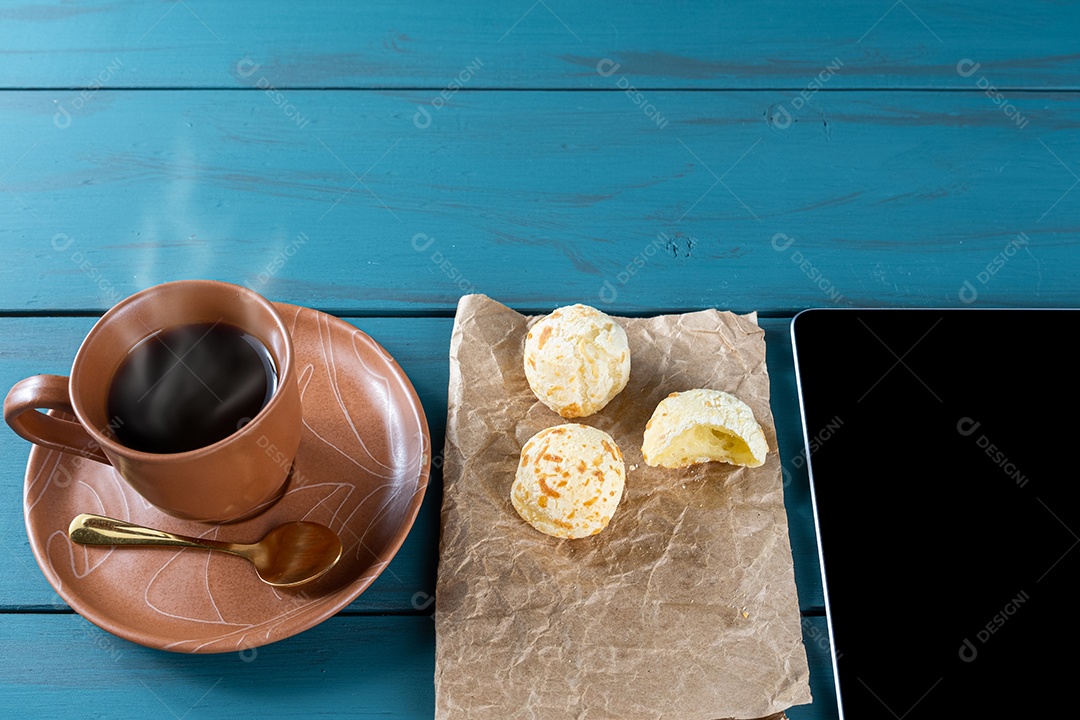 Closeup de pães de queijo brasileiros em um papel pardo, ao lado de tablet, xícara de vista.