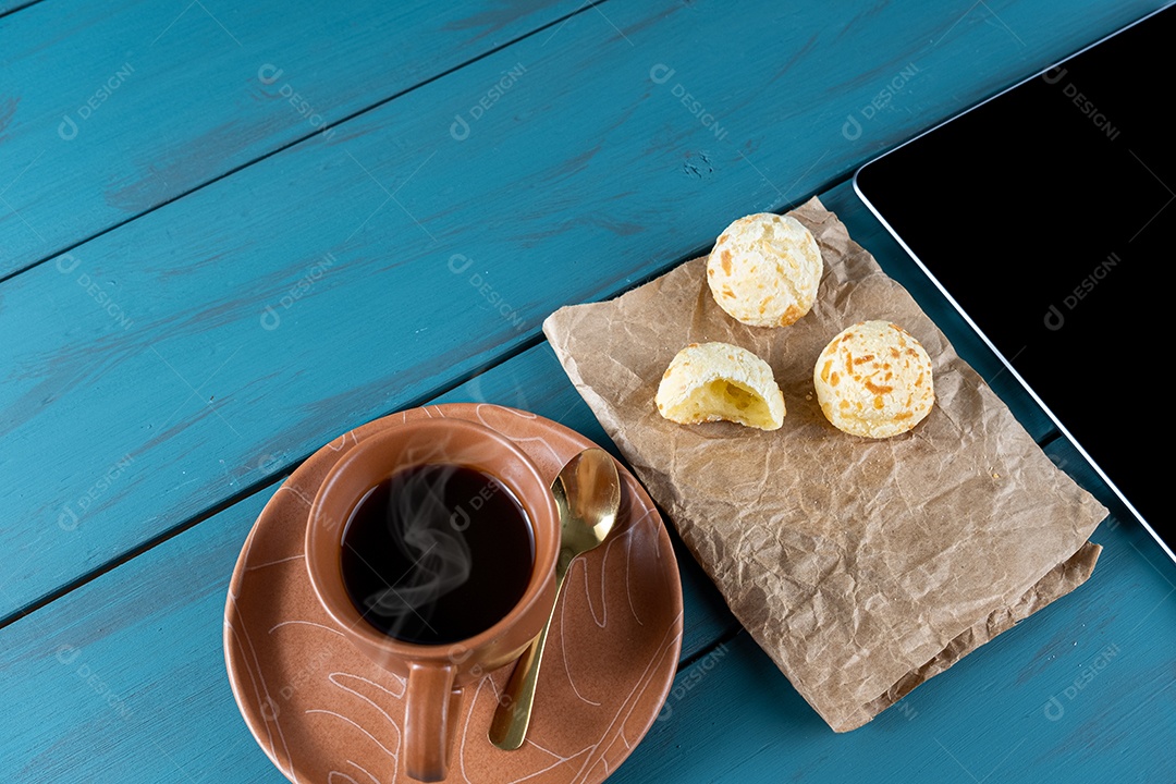 Closeup of Brazilian cheese breads on brown paper next to tablet, cup view.