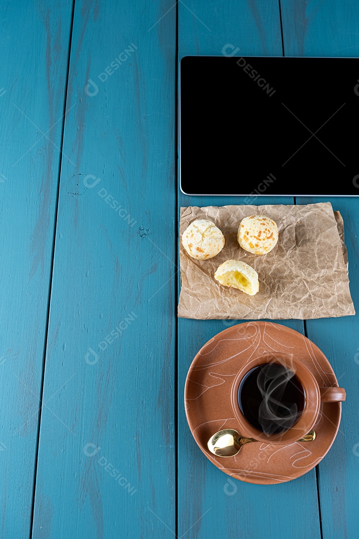 Closeup de pães de queijo brasileiros em um papel pardo, ao lado de tablet, xícara de vista.