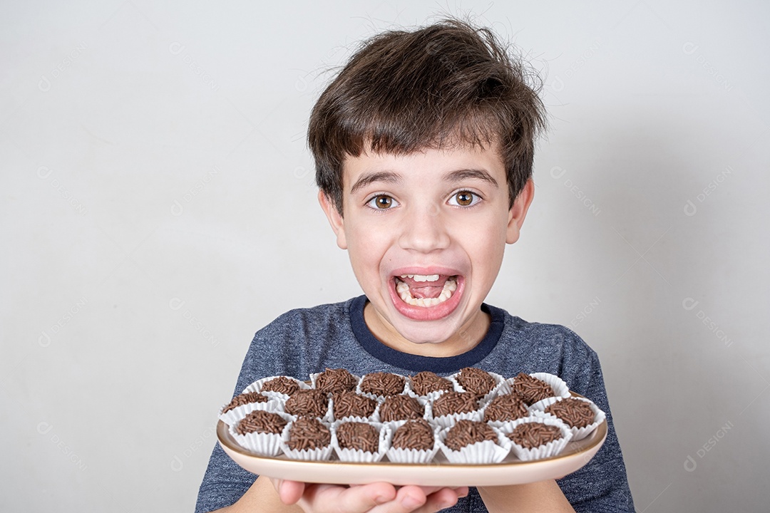 Brasileiro de 9 anos segurando uma bandeja com vários bombons brasileiros e encarando a câmera com um grande sorriso.