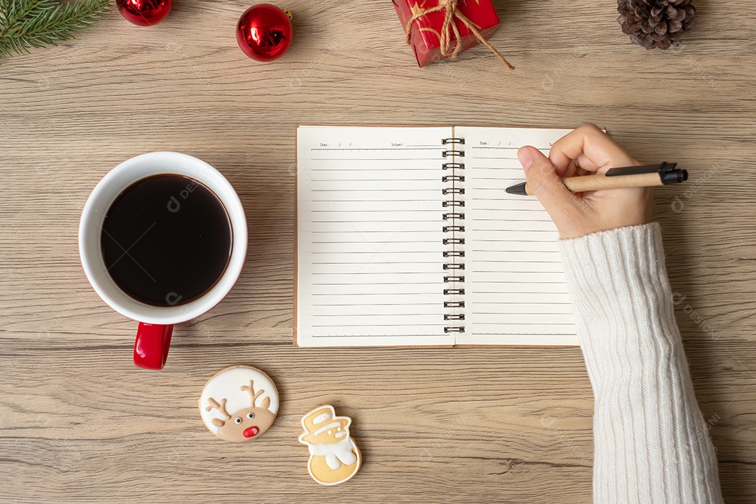 Mão de uma mulher escrevendo no notebook com uma xícara de café preta e Natal