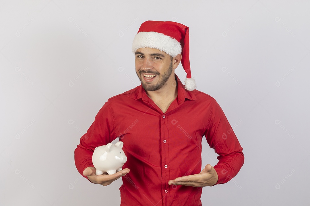 Homem jovem usando gorro Papai Noel segurando cofrinho branco sobre fundo branco