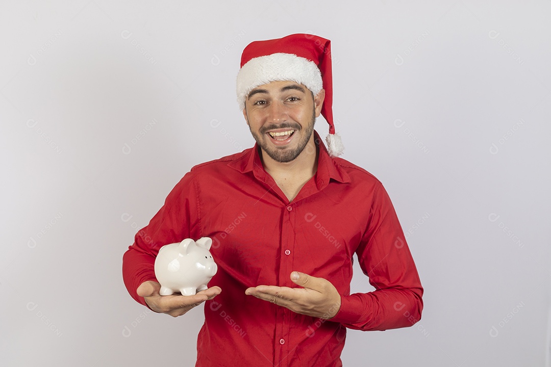 Homem jovem usando gorro Papai Noel segurando cofrinho branco sobre fundo branco