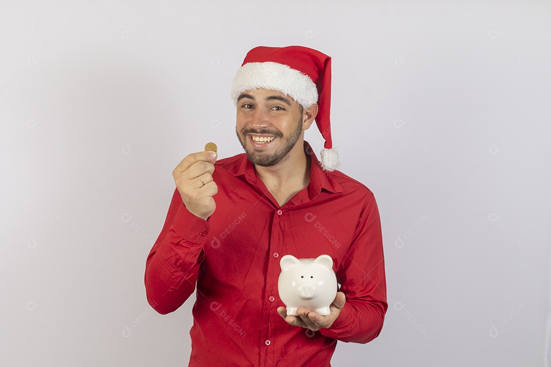 Homem jovem usando gorro Papai Noel segurando cofrinho branco sobre fundo branco