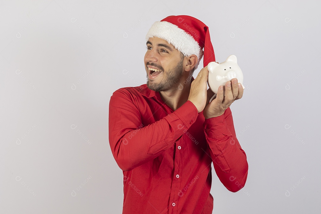Homem jovem usando gorro Papai Noel segurando cofrinho branco sobre fundo branco