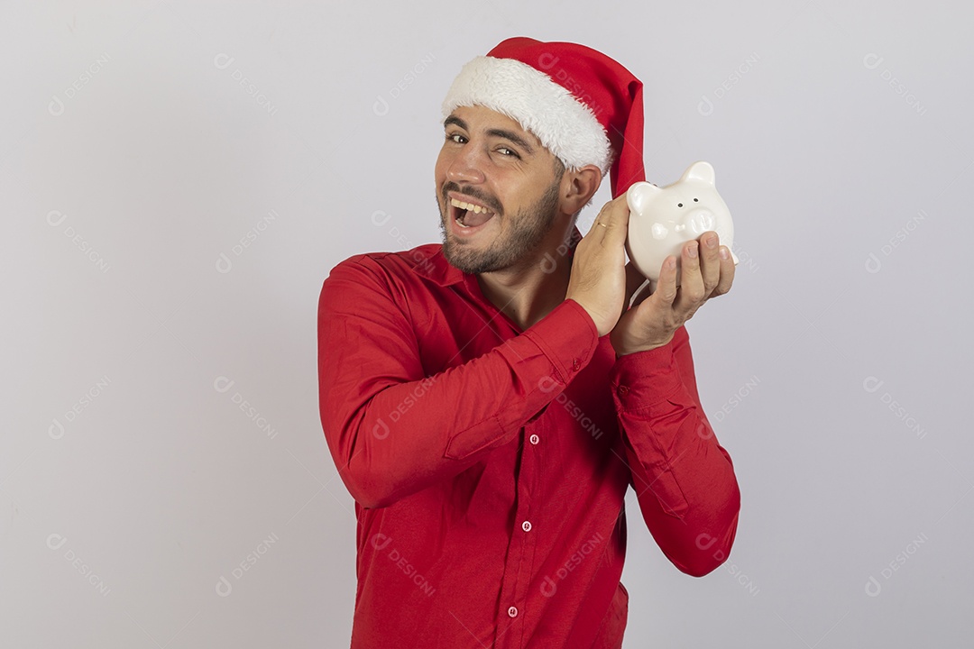 Homem jovem usando gorro Papai Noel segurando cofrinho branco sobre fundo branco