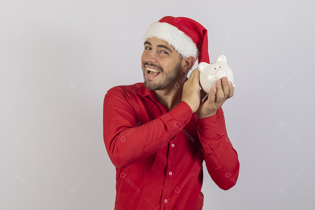 Homem jovem usando gorro Papai Noel segurando cofrinho branco sobre fundo branco