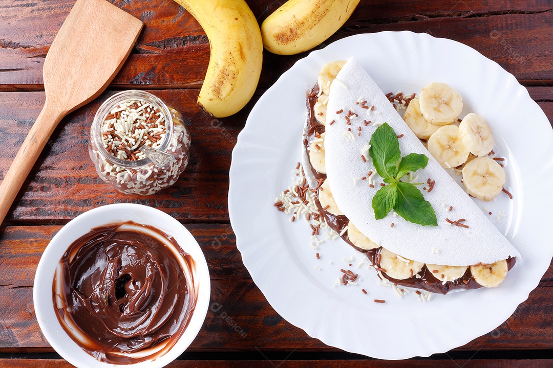 tapioca caseira ou beiju recheado com banana e chocolate na chapa branca sobre mesa de madeira rústica. vista do topo