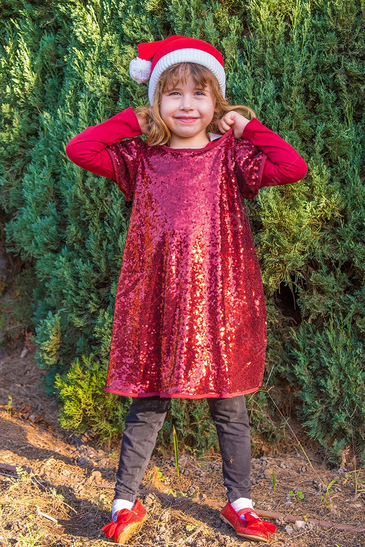 Linda garotinha loira posando em roupas de Natal e fundo de pinheiros.