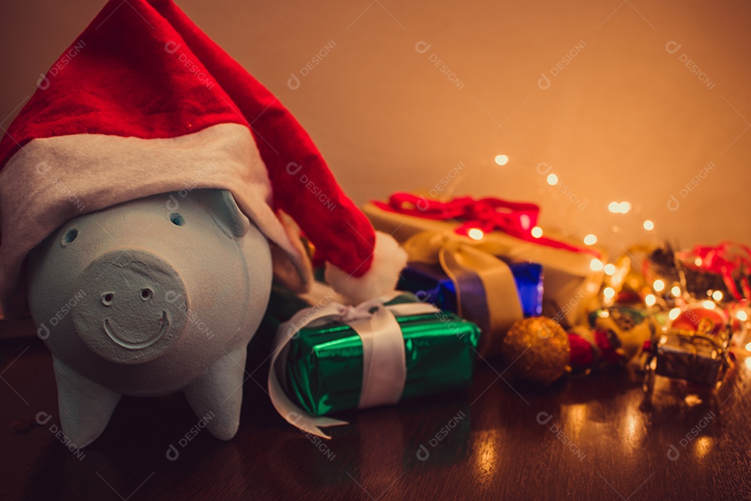 Cofrinho com chapéu de Papai Noel em fundo desfocado de Natal com enfeites, presentes e luz. Espaço para texto.