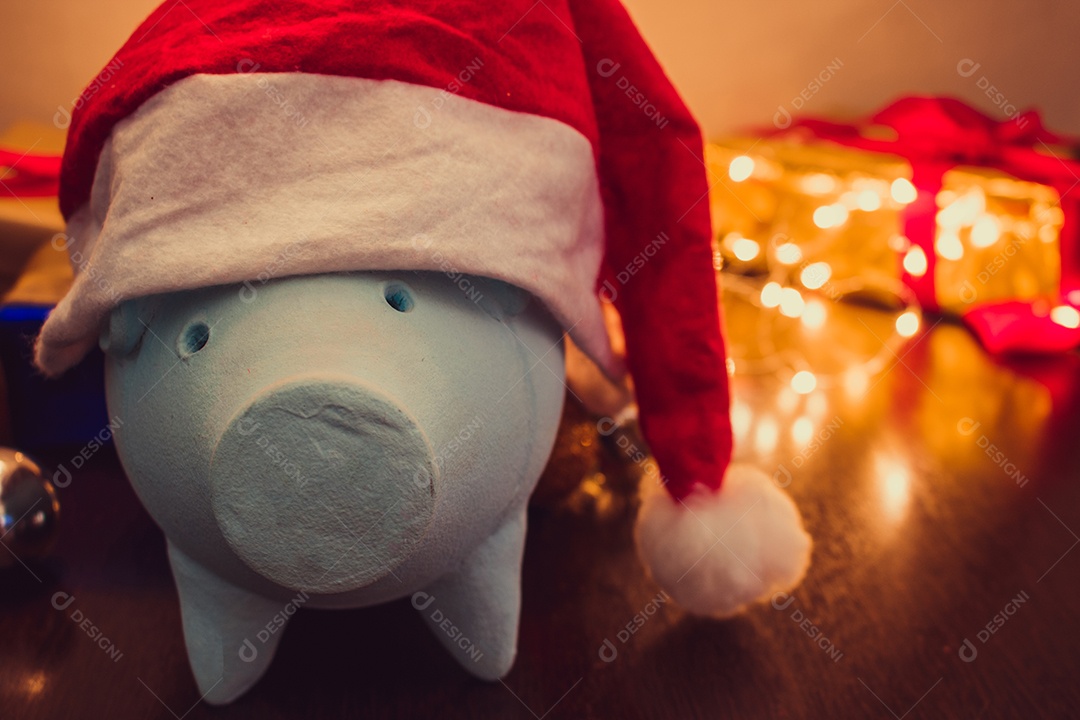 Cofrinho com chapéu de Papai Noel em fundo desfocado de Natal com enfeites, presentes e luz. Espaço para texto.