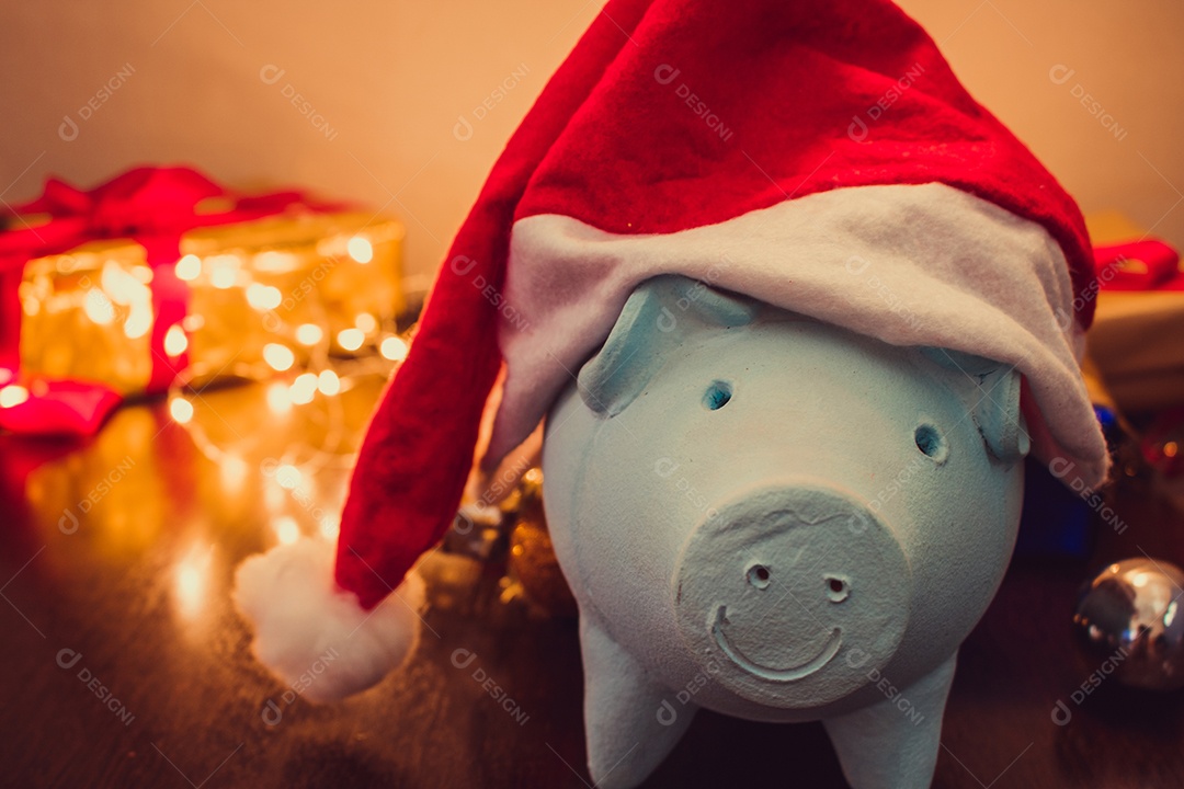 Cofrinho com chapéu de Papai Noel em fundo desfocado de Natal com enfeites, presentes e luz. Espaço para texto.