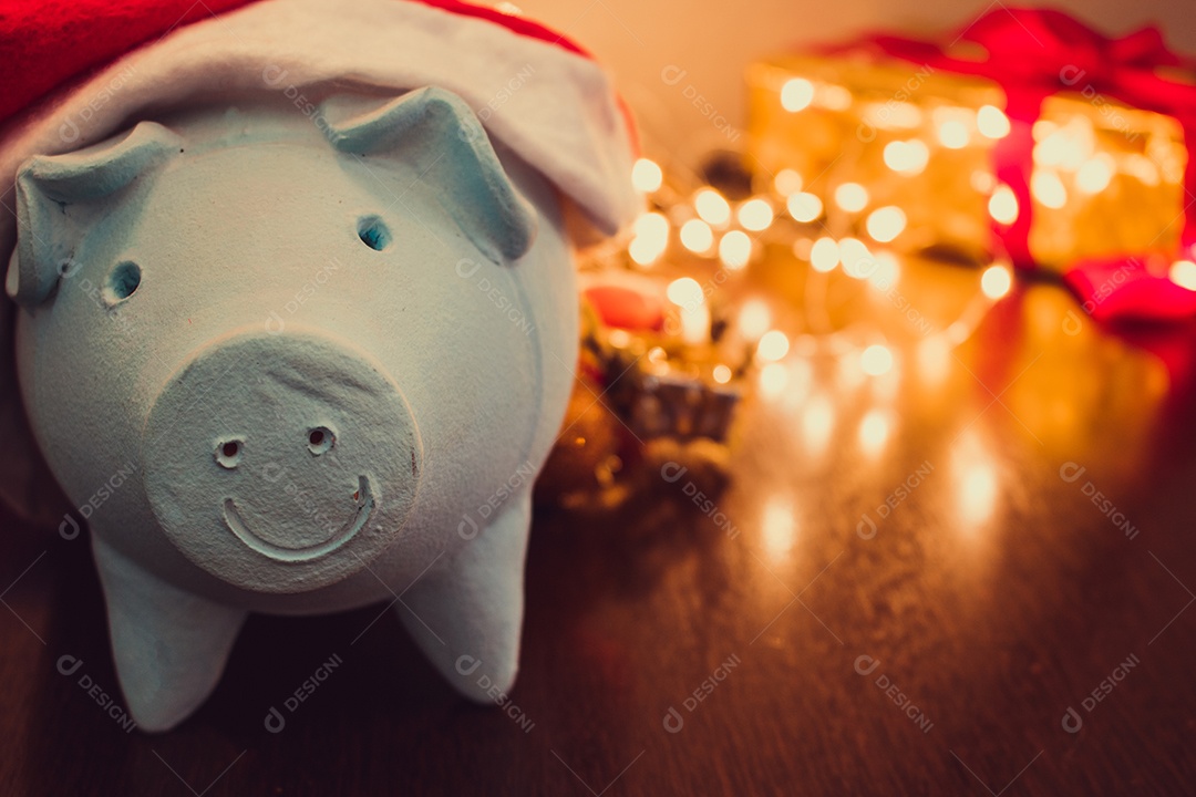 Cofrinho com chapéu de Papai Noel em fundo desfocado de Natal com enfeites, presentes e luz. Espaço para texto.