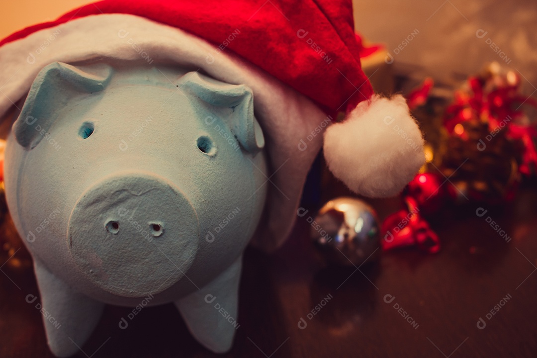 Cofrinho com chapéu de Papai Noel em fundo desfocado de Natal com enfeites, presentes e luz. Espaço para texto.